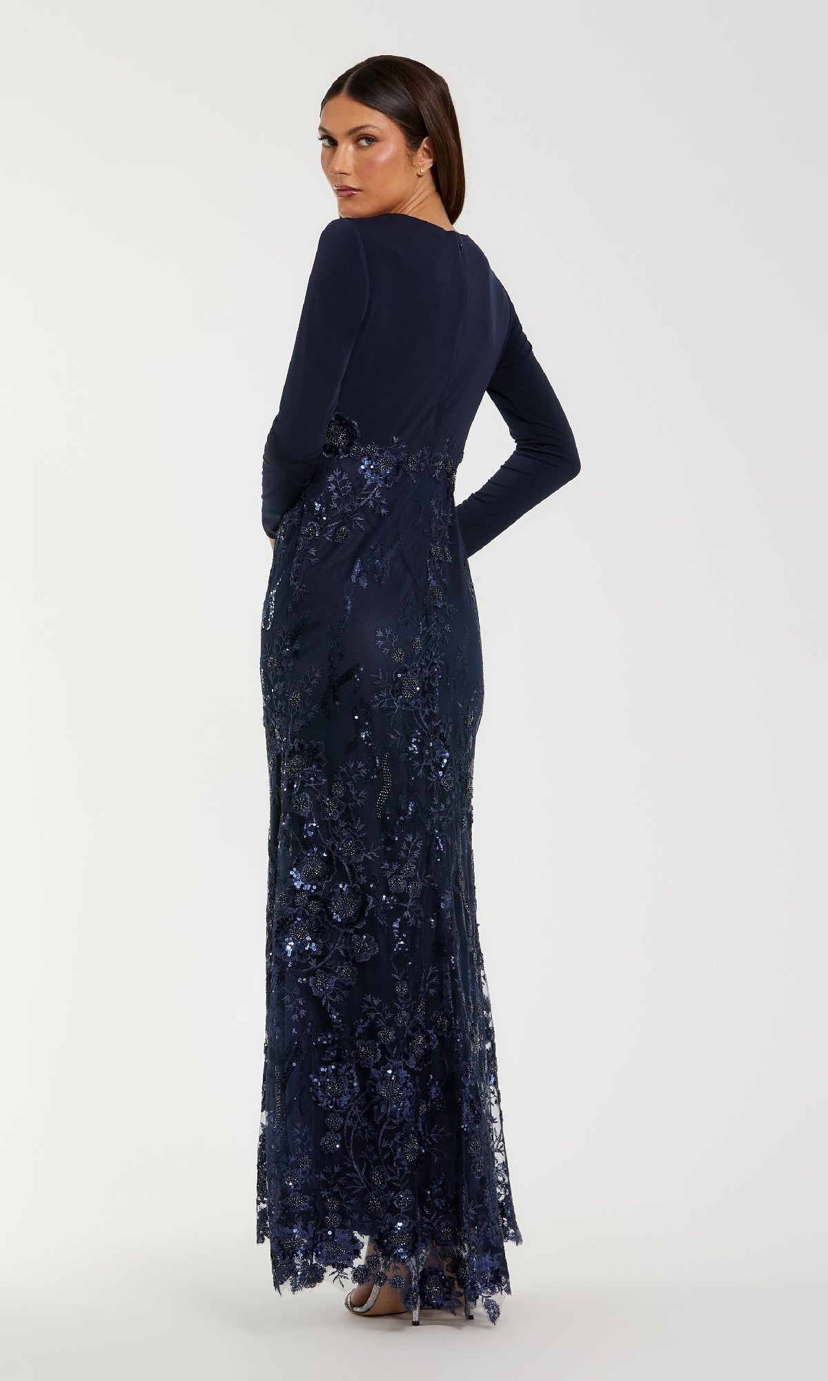 A woman faces away in the Mac Duggal 67871 long sleeve dark blue formal gown, featuring sequins and lace embroidery, paired with silver heels against a plain background.